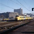 849419, Afbeelding van een trein bestaande uit twee electrische treinstellen mat. 1964 (plan V) van de N.S. bij vertrek uit het N.S.-station Utrecht C.S. te Utrecht. Op de achtergrond het kantoor- en winkelcentrum Hoog Catharijne en een sneltram uit Nieuwegein., Peeters, N.M., fotograaf/schilder